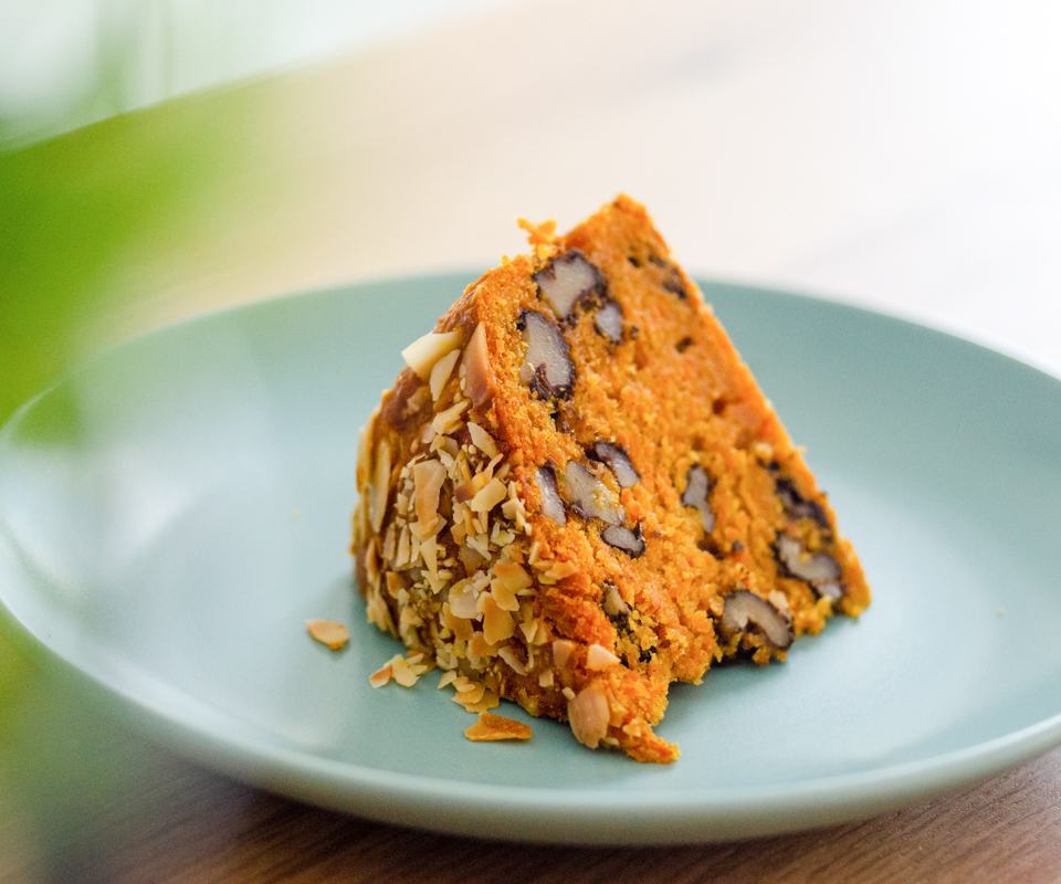 Slice of moist carrot cake with walnuts and almond flakes on top, served on a plate.